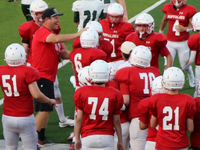 a crowd of young football players gather, with an excited looking coach reaching out and toughing the helmet of one of the players. 