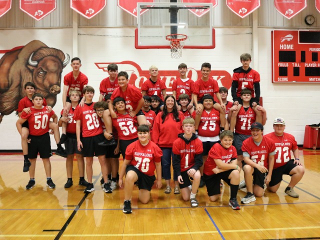a large group of boys dressed in red football jerseys stand around a teacher. 