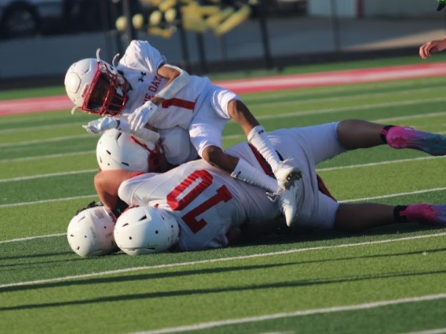 a football player wearing a white jersey that says Lone Oak 1 falls on top of other players on a football field. 