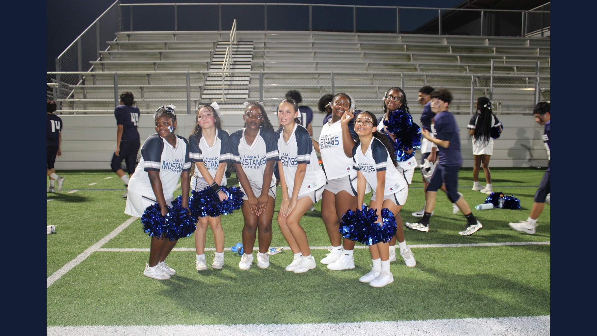 Lamar Mustangs cheerleaders pose together with blue pom-poms on the football field at night.