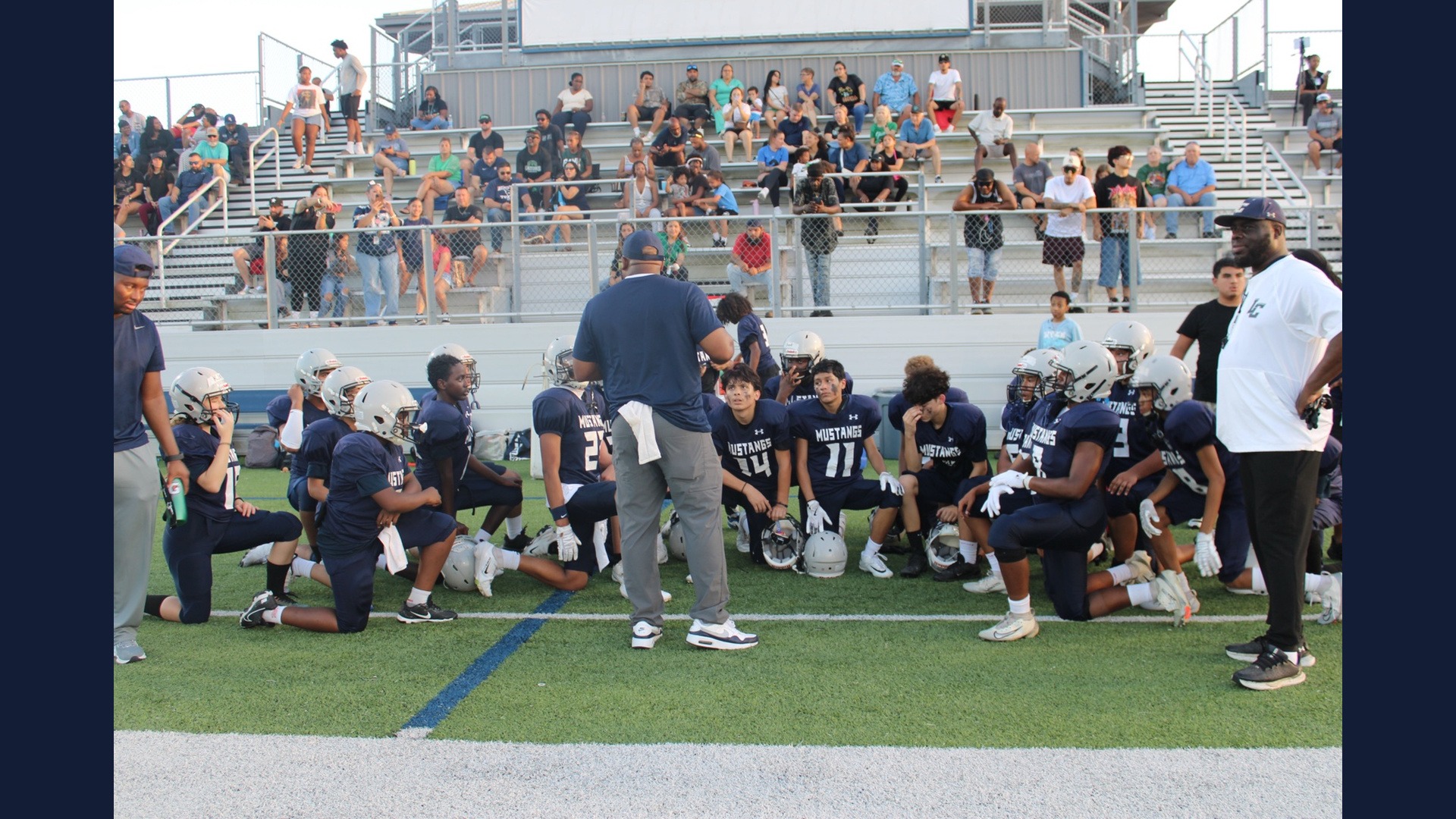 Lamar Mustangs football players gather around their coaches on the field in front of the stands.