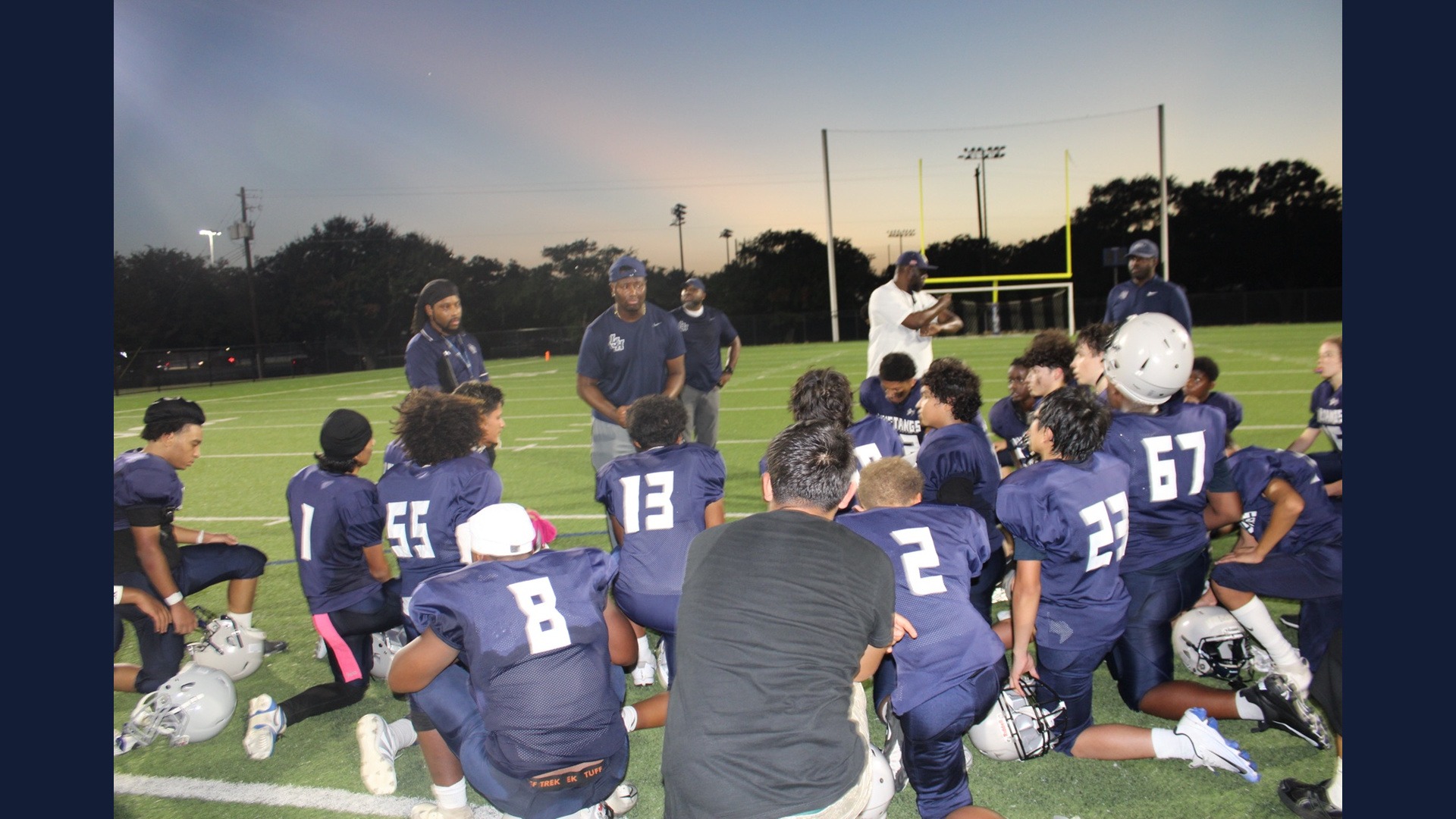 Lamar Mustangs football players gather around their coaches on the field at dusk.
